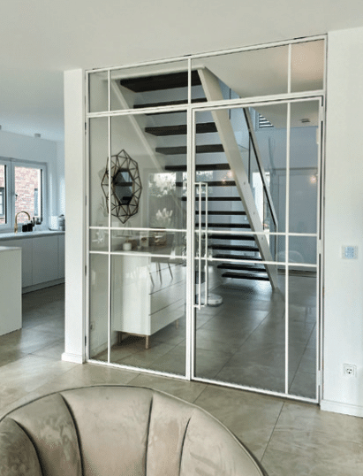 Raumhohe Stahl-Glas-Trennwand mit weißem Rahmen und Drehtür neben offener Treppe